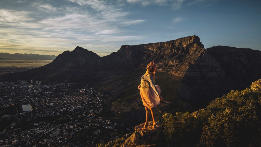 A photograph of me standing on a rock at sunrise, Table Mountain and Devil's peak are behind me. You can see Cape Town City Bowl in the darkness beneath me. 